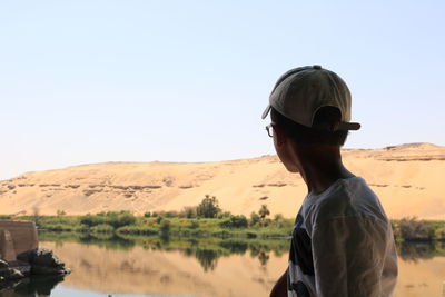 Side view of boy sitting by lake against clear sky