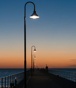Illuminated street lights on pier over sea against clear sky during sunset