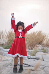 Girl wearing red dress with arms raised standing on rocks against sky