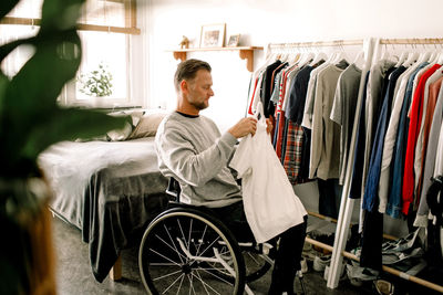 Side view of disabled mature man holding white t-shirt while sitting on wheelchair by clothes rack at home