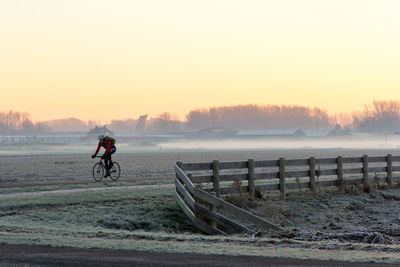 Man riding bicycle on riverbank against sky during sunset