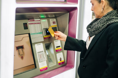 Side view of serious female standing near atm machine with card while buying train ticket while standing in modern terminal