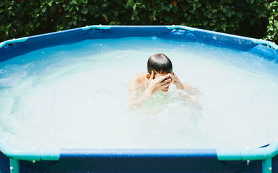 Boy in a pool having a bath removing with his fingers water from face.