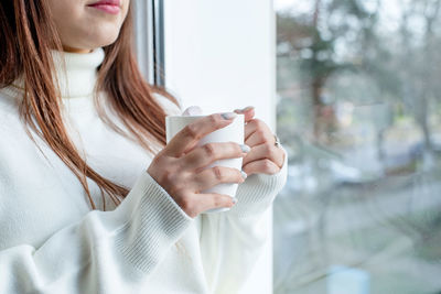Woman in warm white winter sweater sitting on the window at home at christmas eve holding