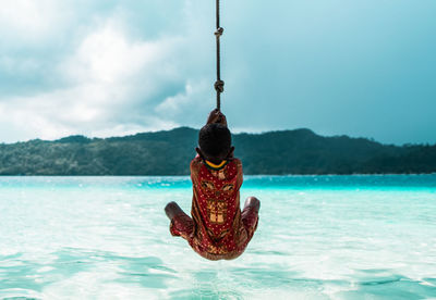 Rear view of boy hanging from rope at beach against sky