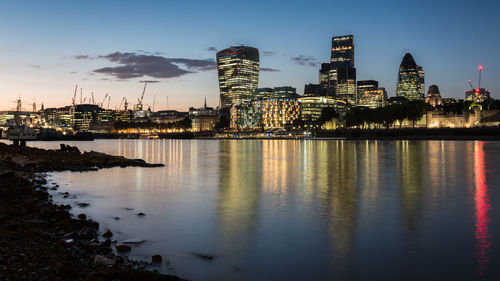 Illuminated modern buildings by thames river against sky in city at dusk