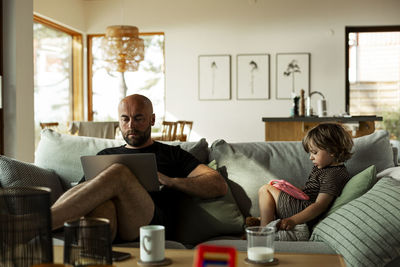 Mature man working over laptop by son using digital tablet sitting on sofa in living room