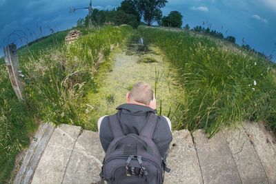 Rear view of man sitting on footbridge over stream at field