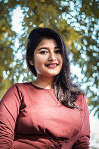 Low angle portrait of smiling young woman standing against trees