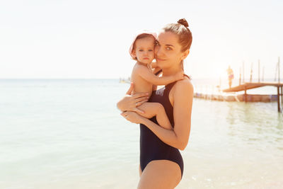 Portrait of smiling mother carrying cute daughter while standing in sea against clear sky