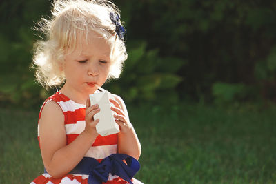 Pretty curly little girl seating in park and drinking milk or juice from box with straw.