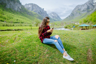 Woman using phone while sitting on grassy field against mountains