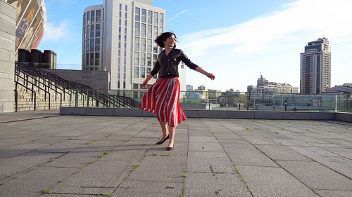 Happy woman wearing striped skirt and jacket on footpath in city