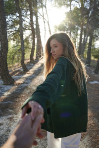 Back view of young long haired female in warm jacket holding hand of anonymous boyfriend while walking together in sunny autumn forest