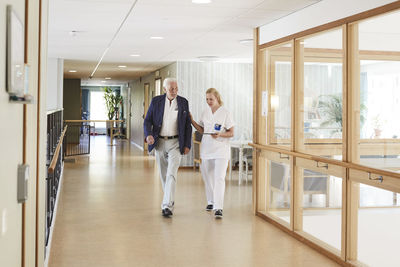 Full length of female nurse and senior male patient walking in hospital corridor