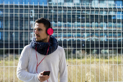 Man wearing headphones looking away standing against fence