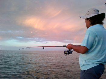 Side view of man fishing in sea against sky during sunset