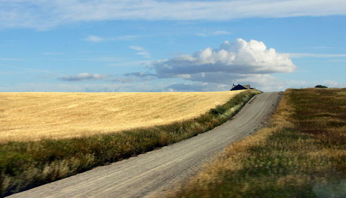 Dirt road passing through field against cloudy sky