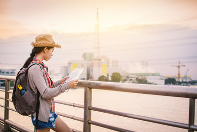 Woman reading paper by railing at lake against sky