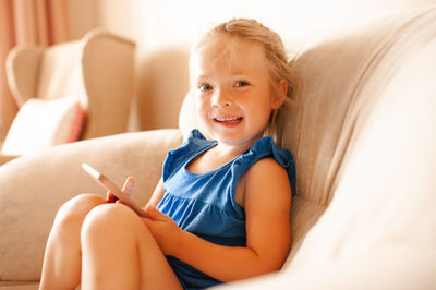 Laughing cute kid girl 3-4 year old holding smart phone watching cartoons sit in armchair in room 