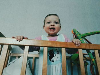 Low angle view of happy baby standing in wooden crib against wall at home