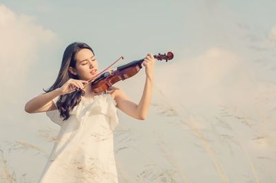 Young woman playing violin while standing against sky