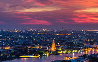 Illuminated city by river against sky at sunset