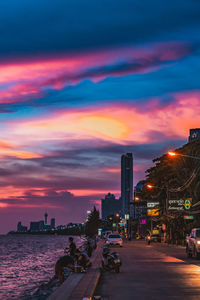 Illuminated road by buildings against sky during sunset