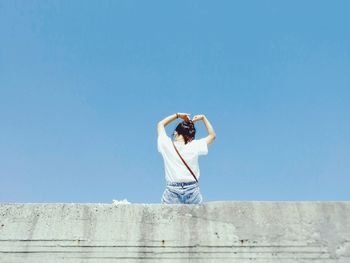 Rear view of young woman sitting on retaining wall against clear blue sky