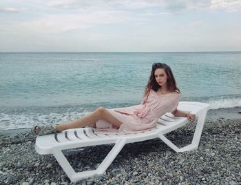 Young woman sitting on chair at beach against sky