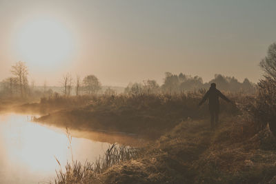 Rear view of silhouette man standing by lake against sky at sunrise