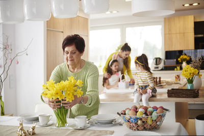 Senior woman arranging flowers on dining table with family in background