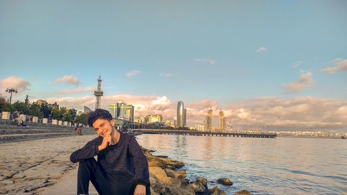 Portrait of young man sitting at riverbank against sky in city