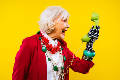 Senior woman wearing sunglasses screaming on telephone standing against yellow background