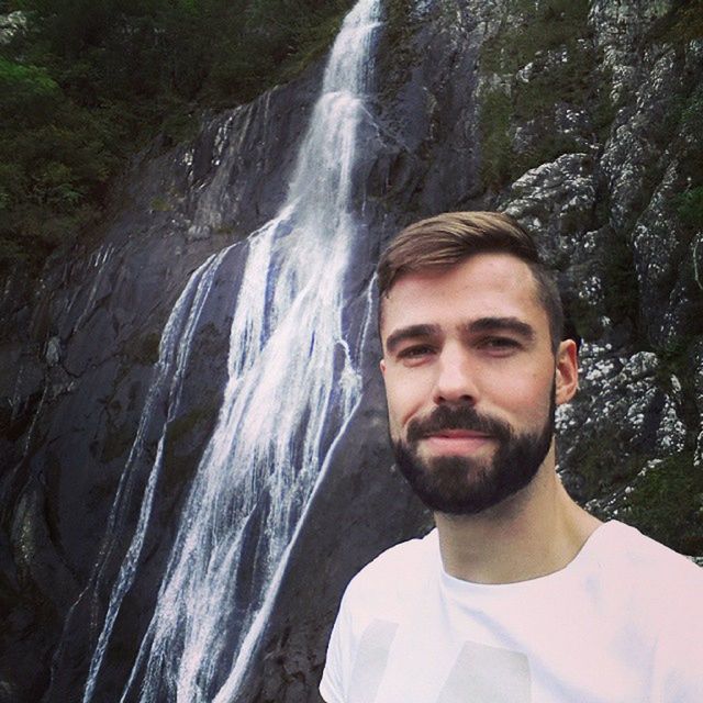 lifestyles, leisure activity, water, portrait, looking at camera, waterfall, person, young adult, rock - object, young men, front view, flowing water, nature, smiling, motion, casual clothing, forest, standing