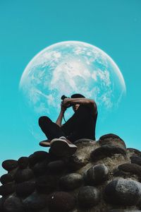 Low angle view of man sitting on rock against moon in blue sky