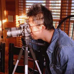 Side view of man photographing with camera against window blinds