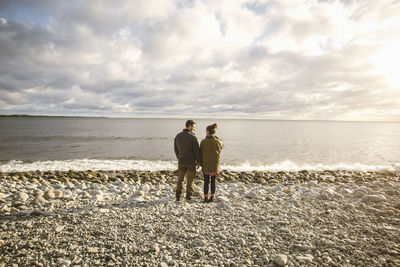 Rear view of couple standing on rocky shore at beach against sky