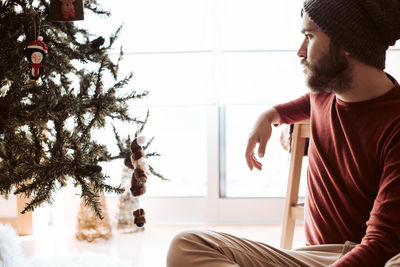 Man looking away while sitting by christmas tree at home