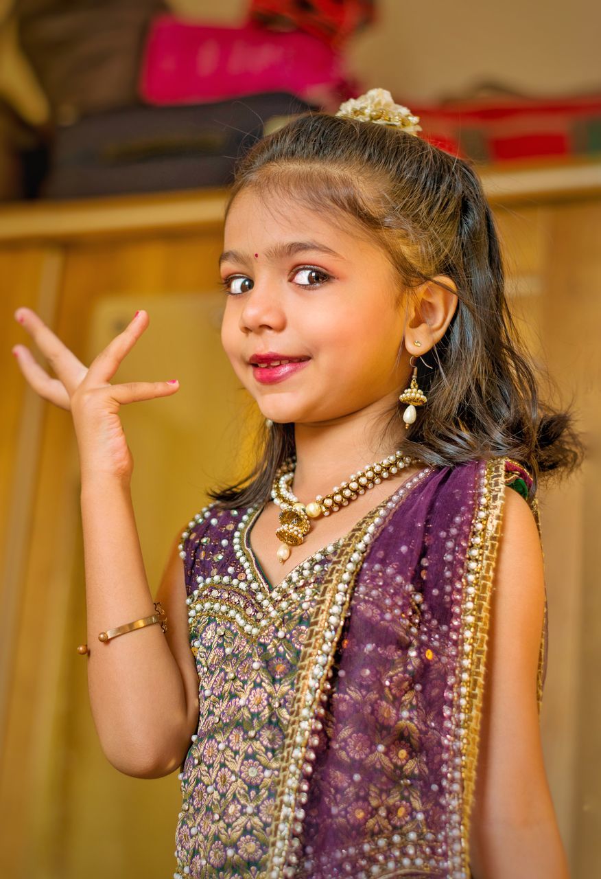 one person, women, smiling, child, childhood, happiness, portrait, female, indoors, emotion, waist up, looking at camera, cheerful, clothing, fashion, dress, cute, yellow, jewelry, traditional clothing, celebration, pink, standing, arts culture and entertainment, focus on foreground, hairstyle, lifestyles, person, brown hair, innocence, fashion accessory, human face, photo shoot, hand, teeth, front view