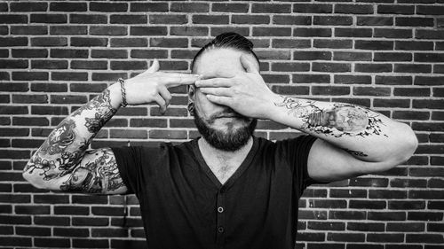 Young man with tattoo on hands covering eyes against brick wall