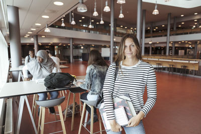 Portrait of female university student standing at cafeteria with friends in background