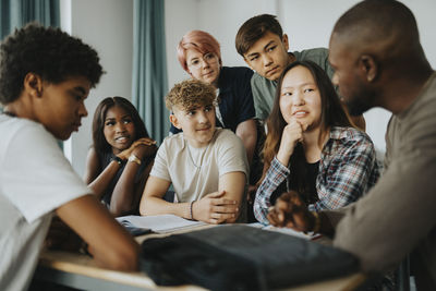 Teenage girls and boys with non-binary person looking at teacher discussing in classroom