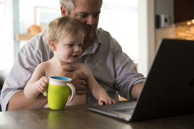 Dad working from home on computer holds toddler with green sippy cup