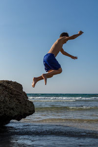 Full length side view of shirtless boy jumping from rock into sea