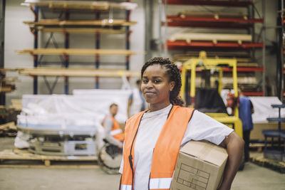 Portrait of smiling female carpenter carrying cardboard box in warehouse