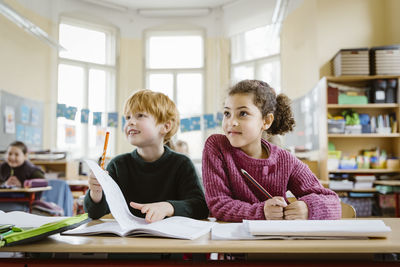 Smiling blond boy sitting with girl at desk looking away in classroom