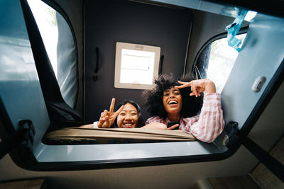 From below of cheerful young diverse female friends showing peace sign and looking at camera through window inside camper van while having fun together during summer vacation