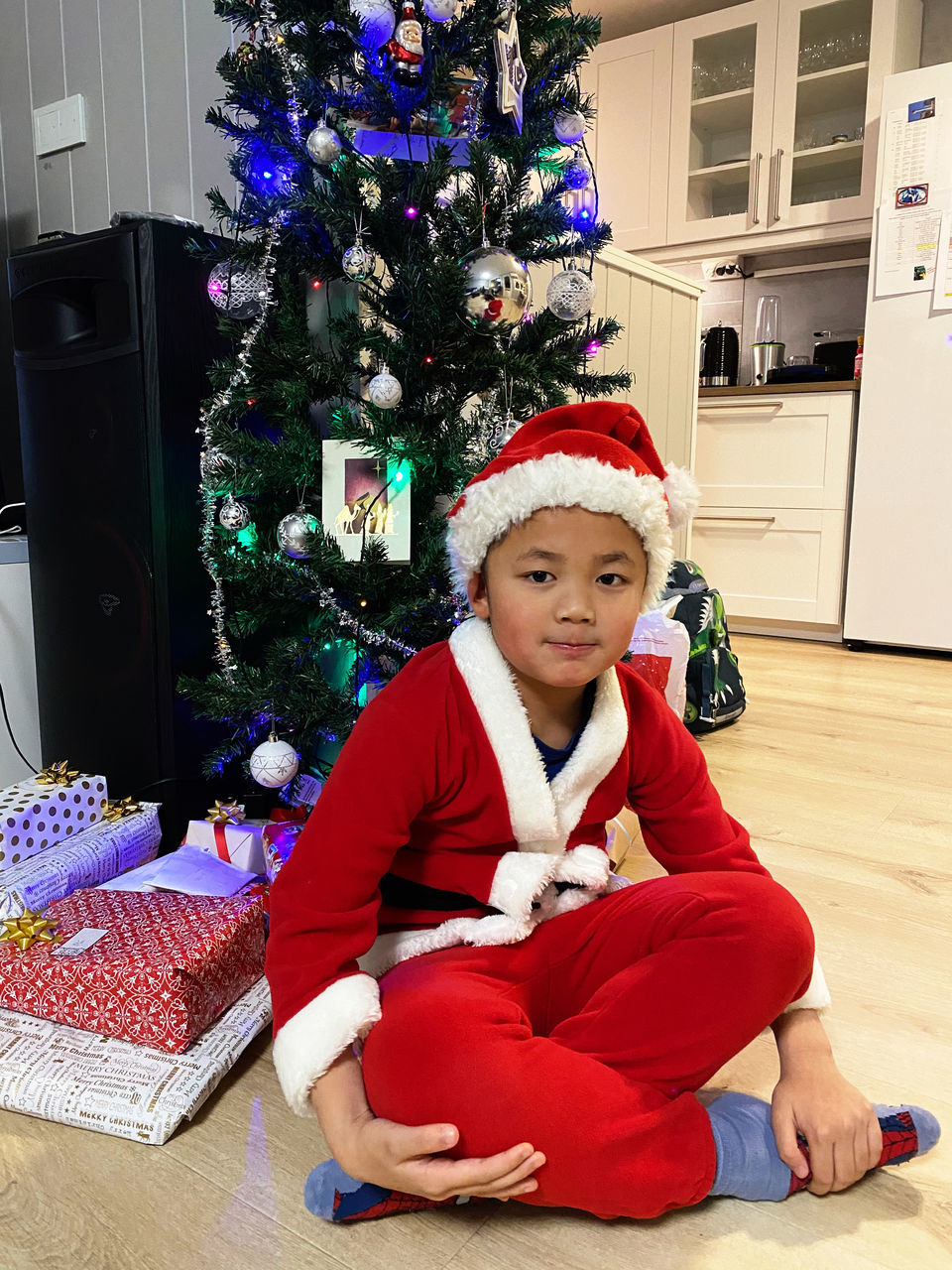 christmas, celebration, holiday, child, childhood, christmas tree, one person, real people, sitting, holiday - event, indoors, full length, front view, emotion, gift, men, males, cute, santa hat, innocence, flooring, christmas ornament