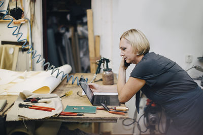 Side view of mature female upholstery worker looking at laptop while talking on phone in workshop